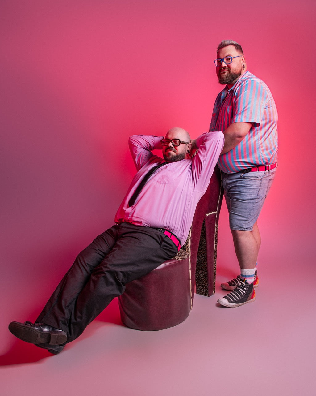 Two men posing against a pink background, one sitting on a round stool and the other standing.