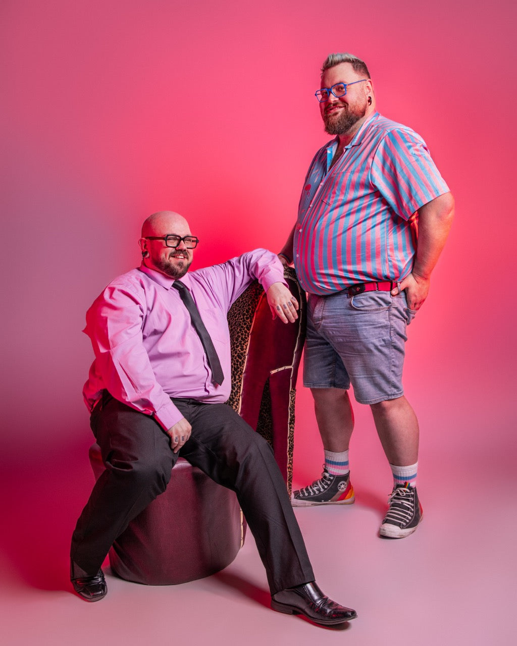 Two men posing against a pink background, one sitting.