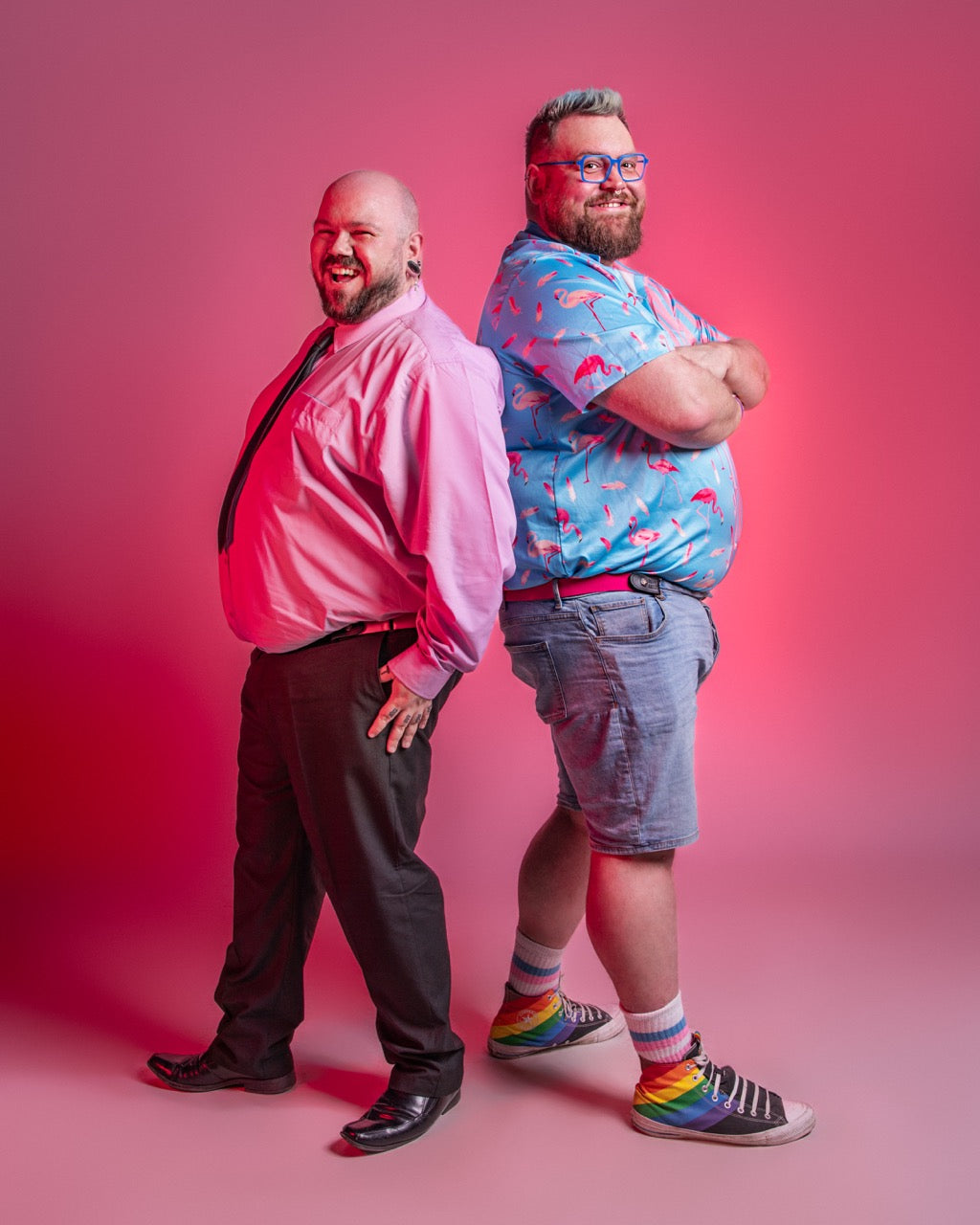Two people standing against a pink background, both wearing pink Bear Belts, one wearing a pink shirt and black pants, the other in a blue patterned shirt and shorts with colorful socks.